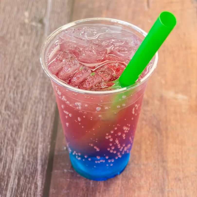 Clear plastic cup of blue and red layered soda with ice and a bright green straw on a wooden table.
