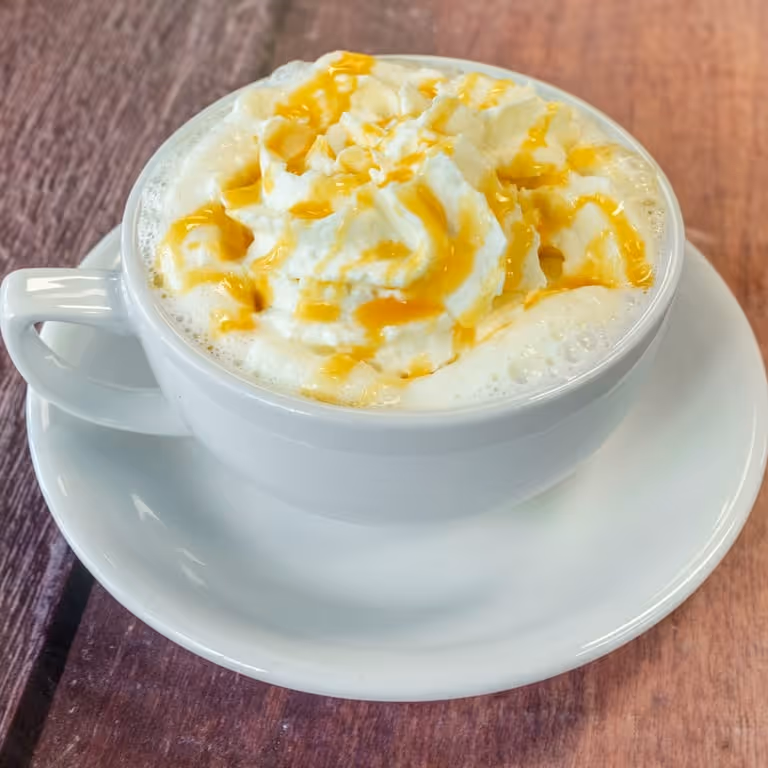 White cup of coffee topped with whipped cream and caramel drizzle on a white saucer over wooden surface.