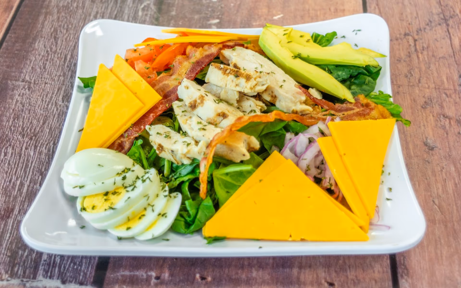 Salad with grilled chicken, cheddar cheese slices, bacon, avocado, hard-boiled egg, and mixed greens on white plate.