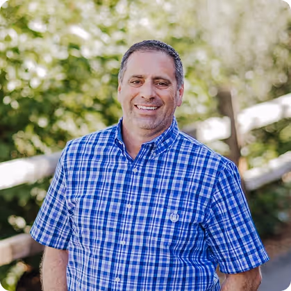 Smiling man in a blue checkered shirt standing outdoors with trees and a fence in the background.