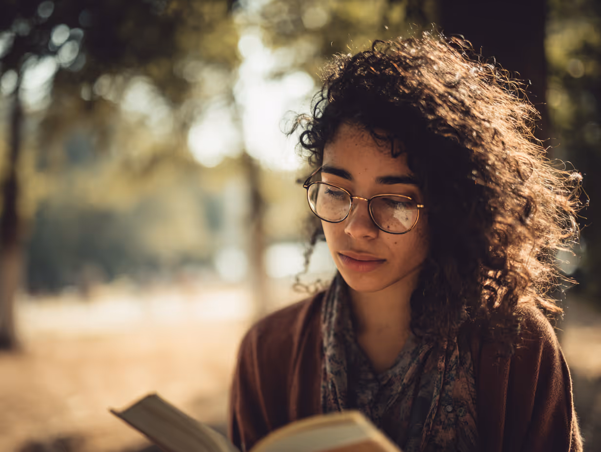 Young woman with curly hair and glasses reading a book outdoors in soft sunlight.