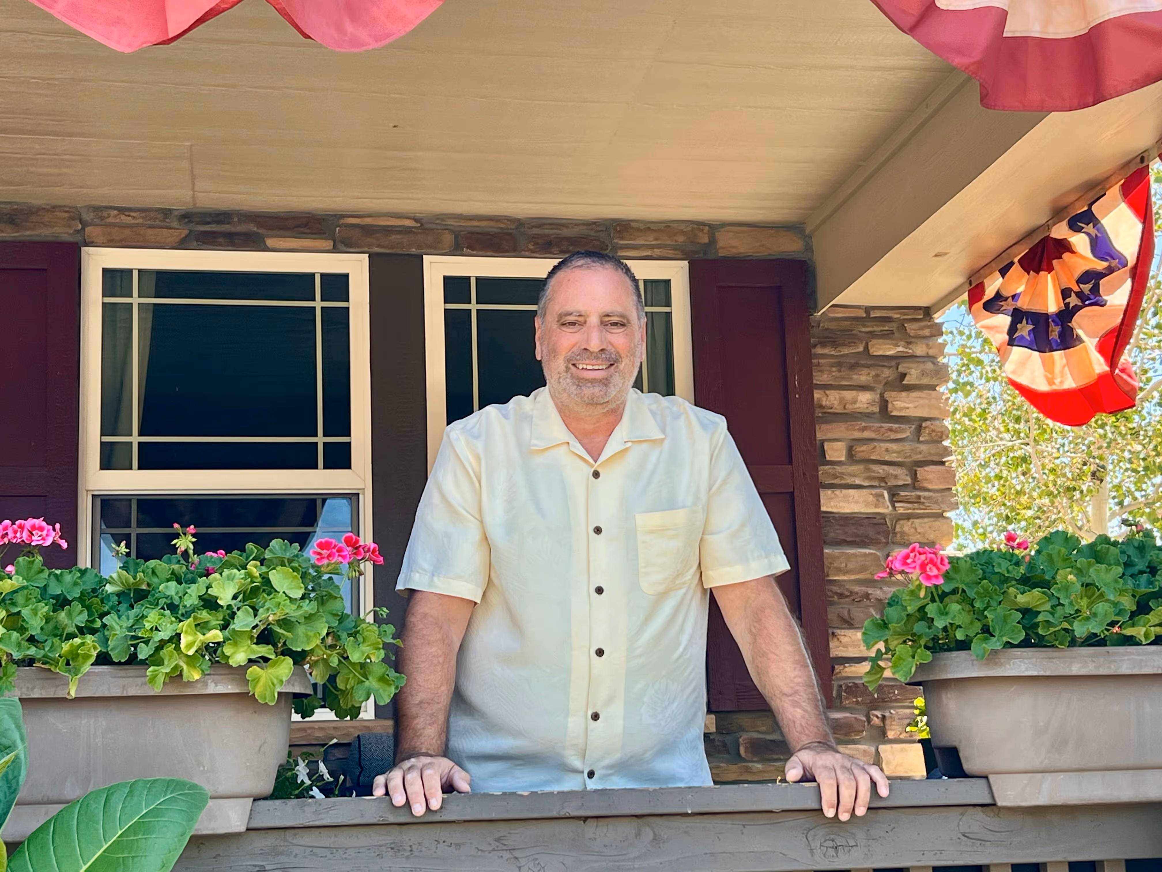 Smiling man in a light yellow short-sleeve button shirt on a porch with flower pots and patriotic bunting.