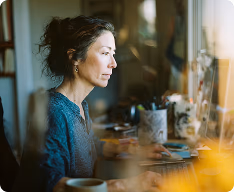 Woman with dark hair focused on computer work at a desk near a window with office supplies.