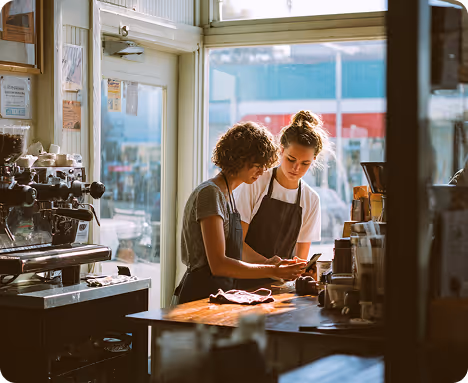 Two baristas in aprons looking at a phone behind a coffee shop counter near a large window.