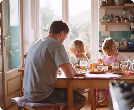 Man and two young girls sitting at a kitchen table eating breakfast with sunlight through window.