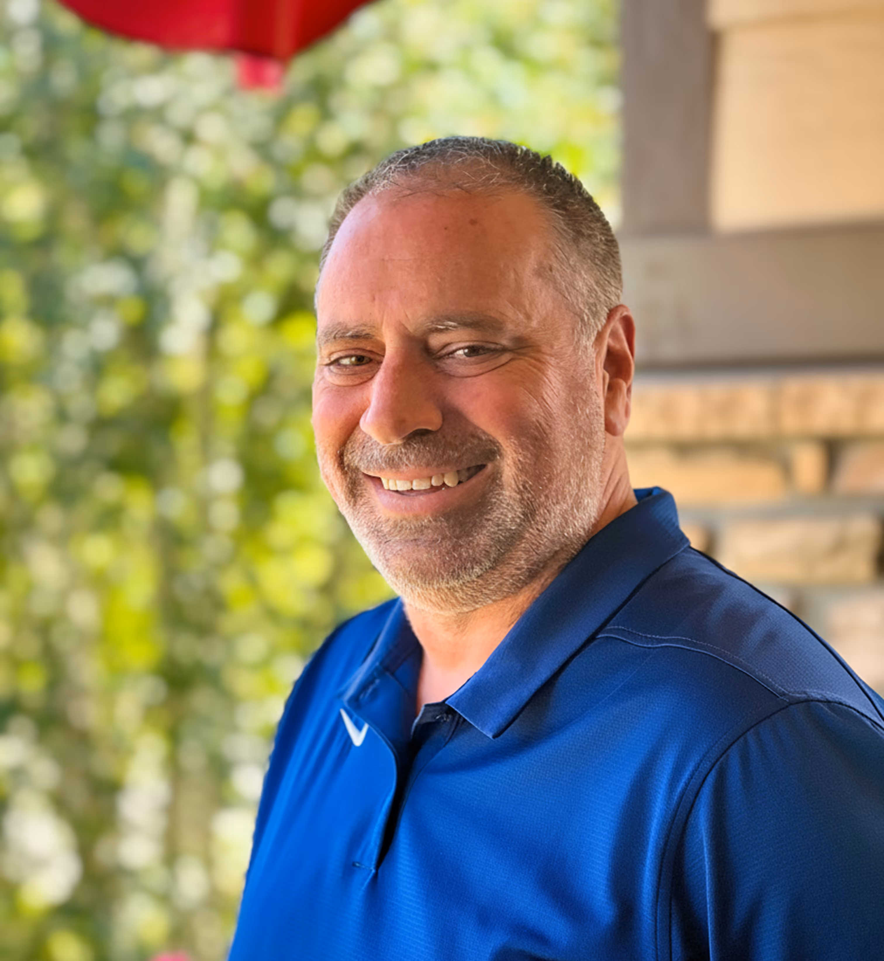 Smiling man with short hair and stubble wearing a blue polo shirt outdoors near foliage.