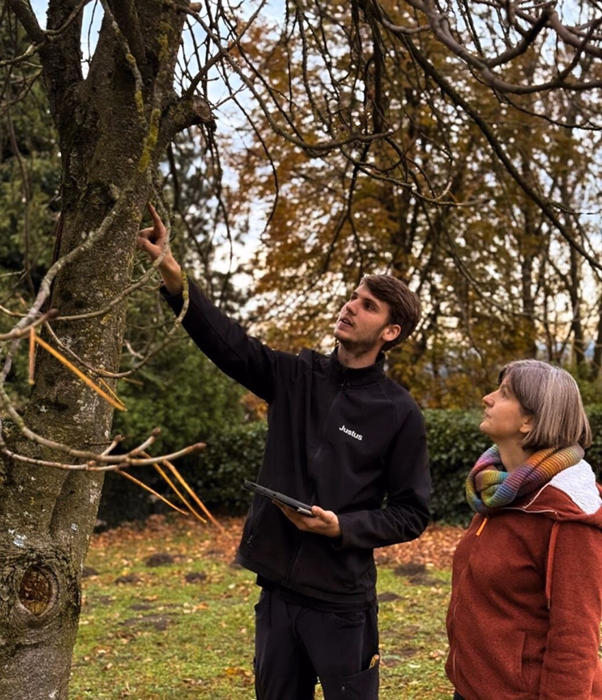Man in black jacket showing tree branches to woman in red coat with rainbow scarf in park.
