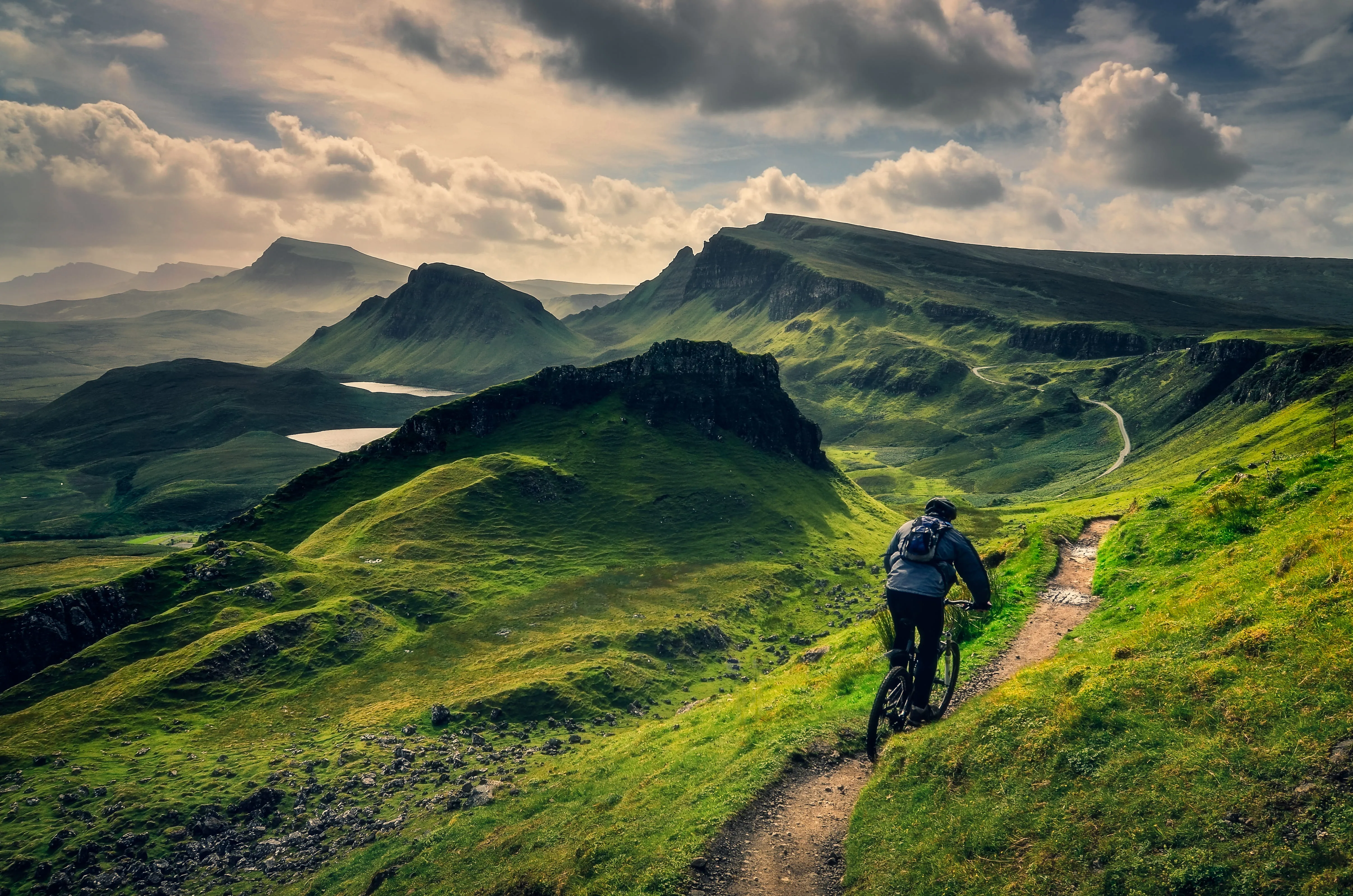 Mountain biker rides on narrow dirt trail through green hills under a cloudy sky.