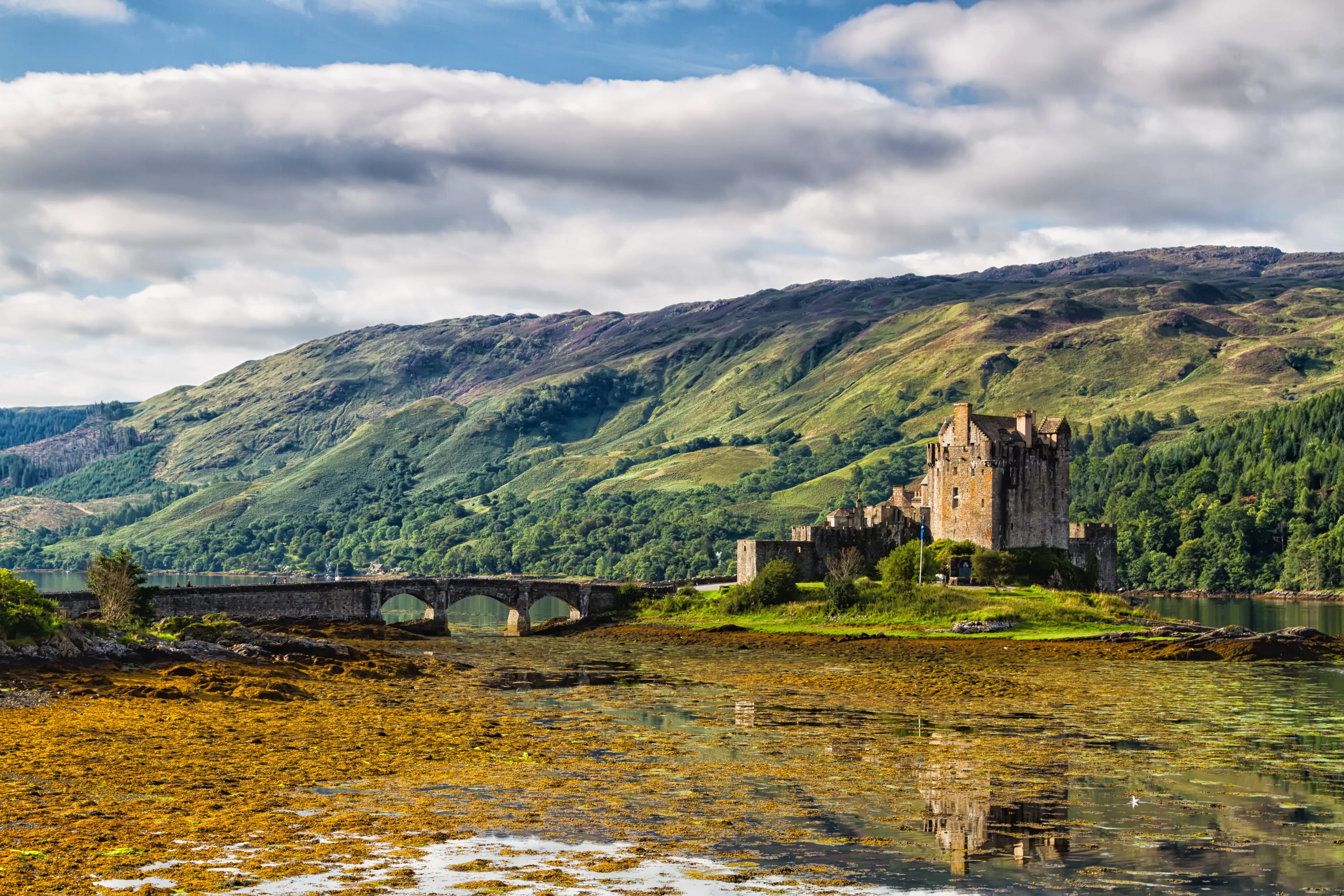 Stone castle on a small island connected by a bridge with lush green hills and cloudy sky behind.