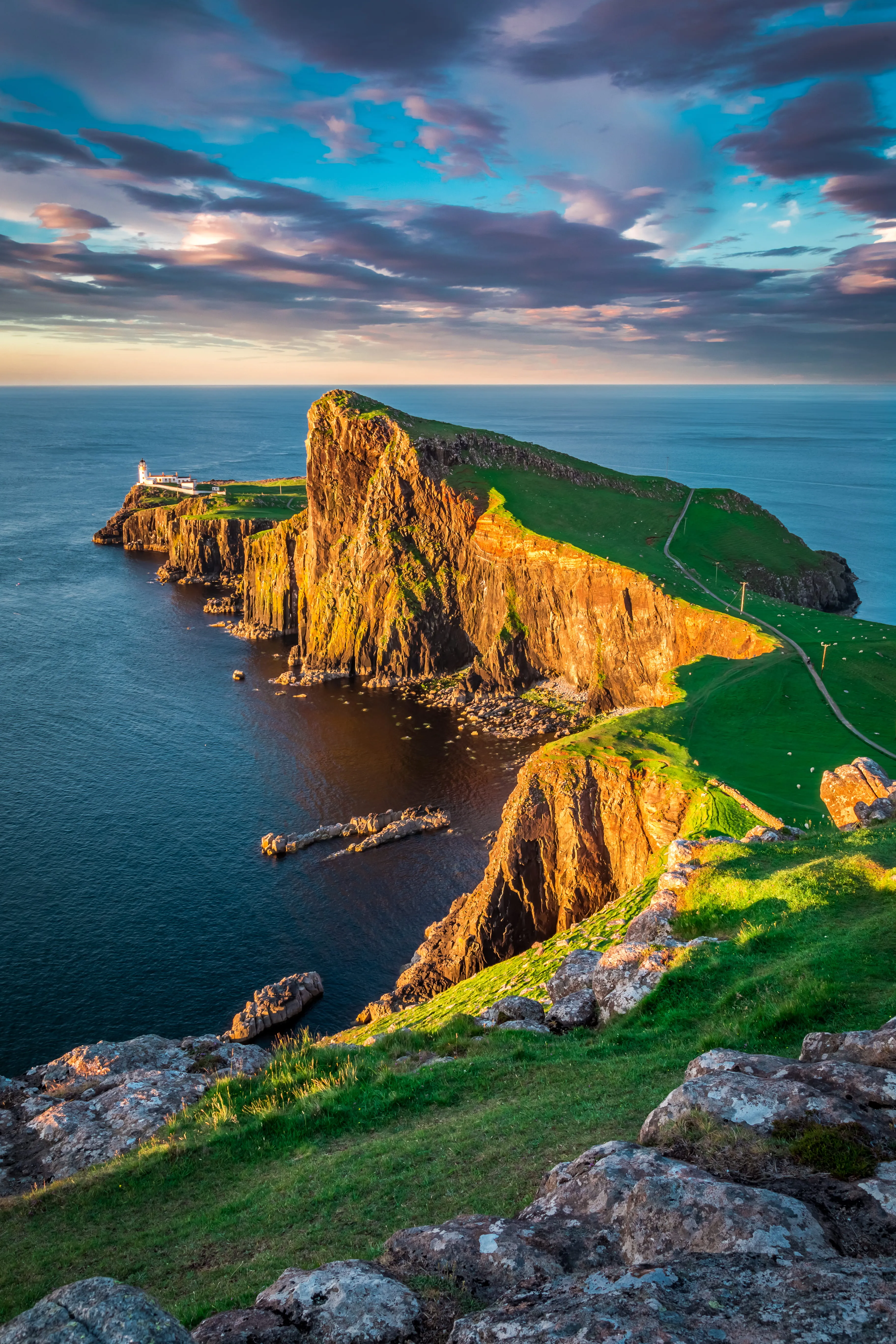 Neist Point lighthouse on rugged cliffs lit by sunset, overlooking calm sea under a partly cloudy sky.
