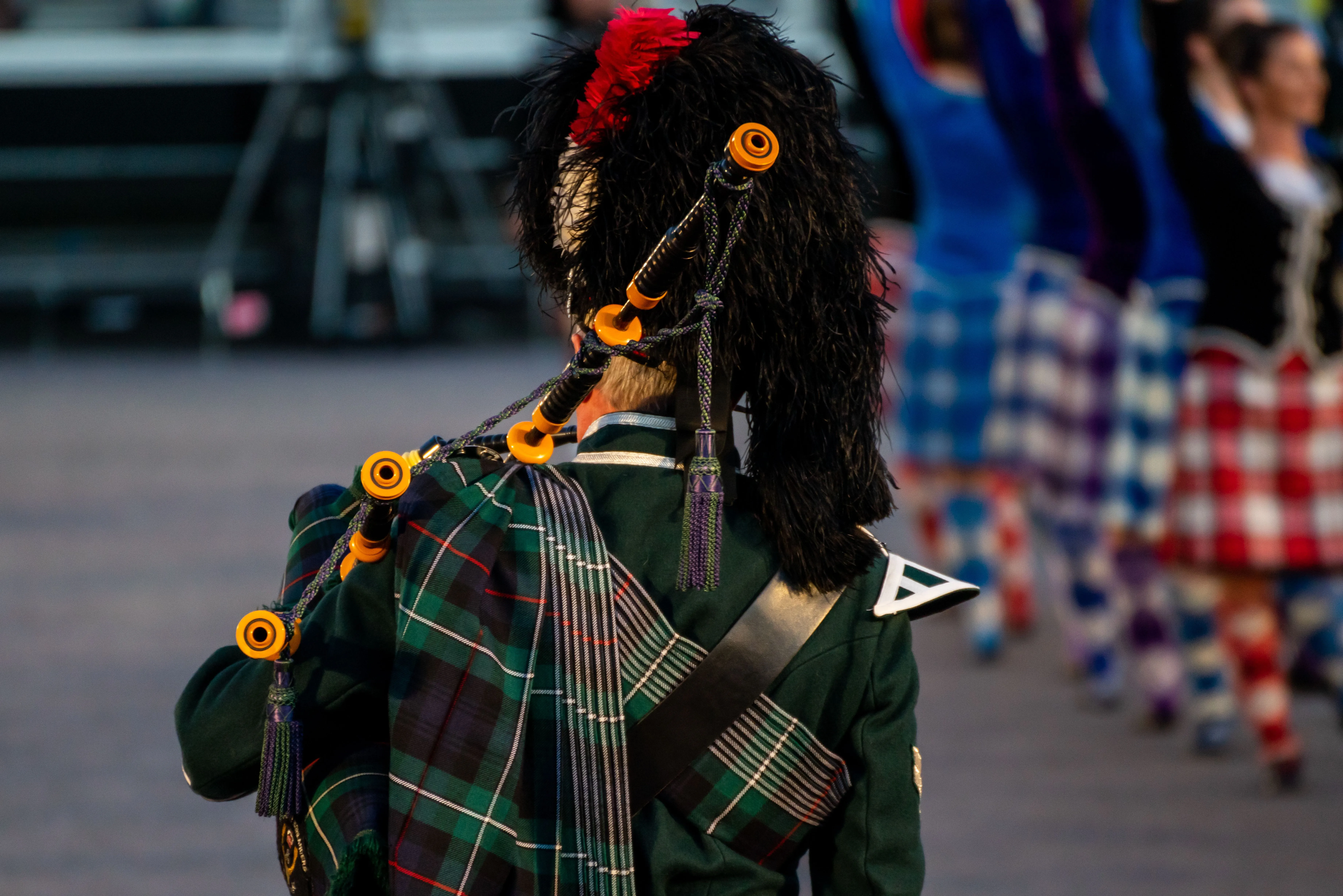Man in traditional Scottish attire playing bagpipes with dancers wearing kilts in the background.
