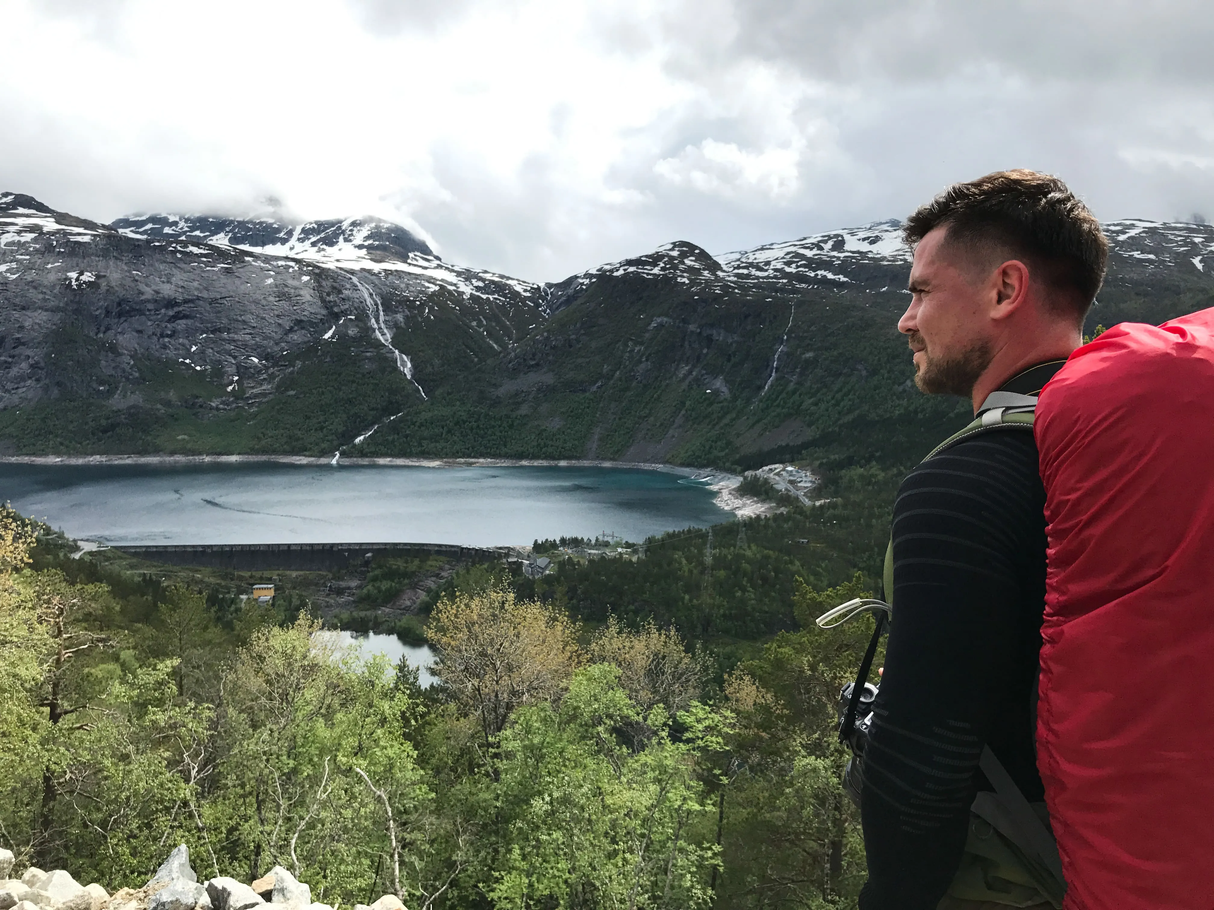Hiker with red backpack overlooking a lake, forest, and snow-capped mountains under cloudy sky.