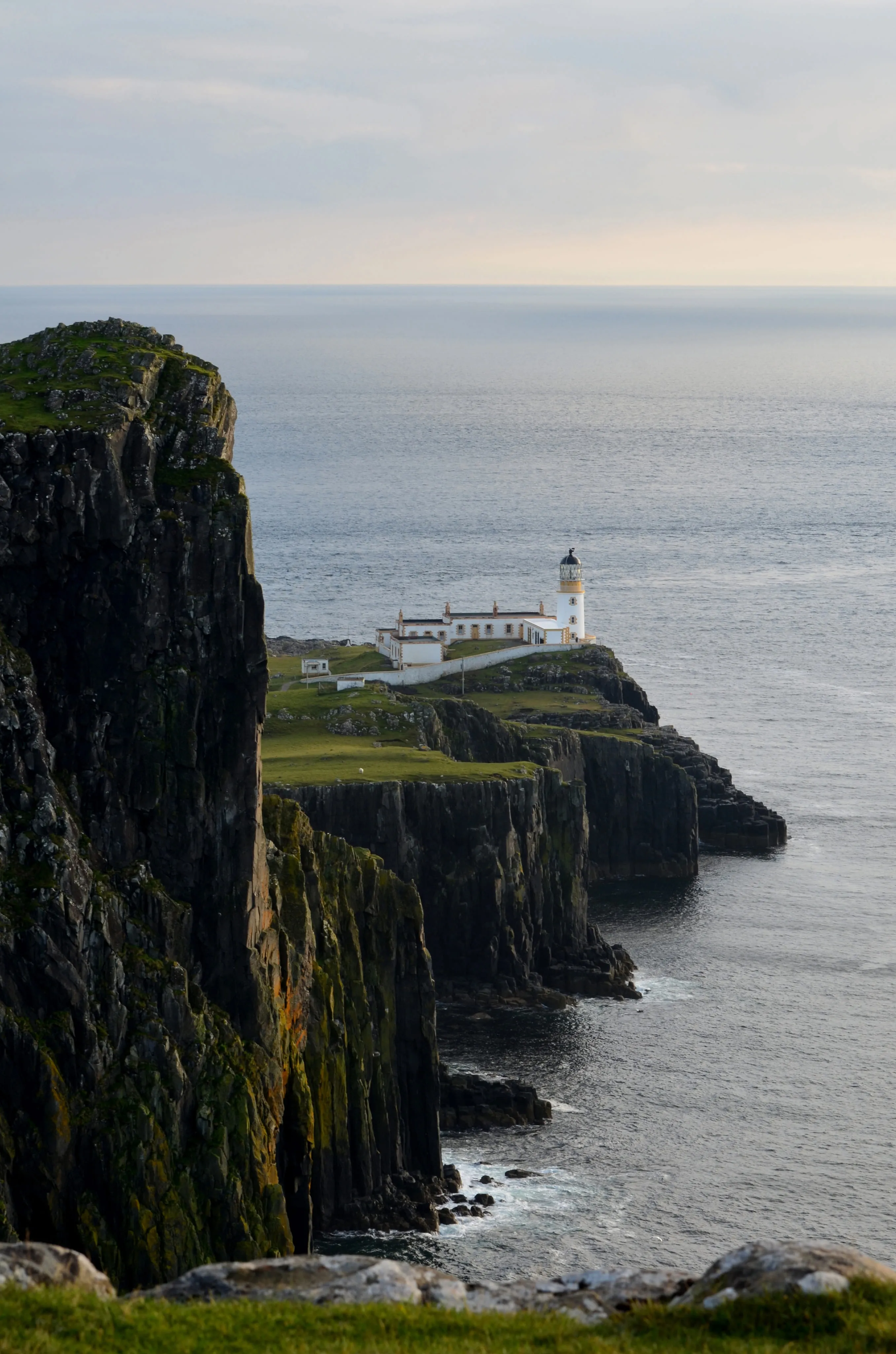 White lighthouse and buildings on green cliffside overlooking a calm sea under a cloudy sky.
