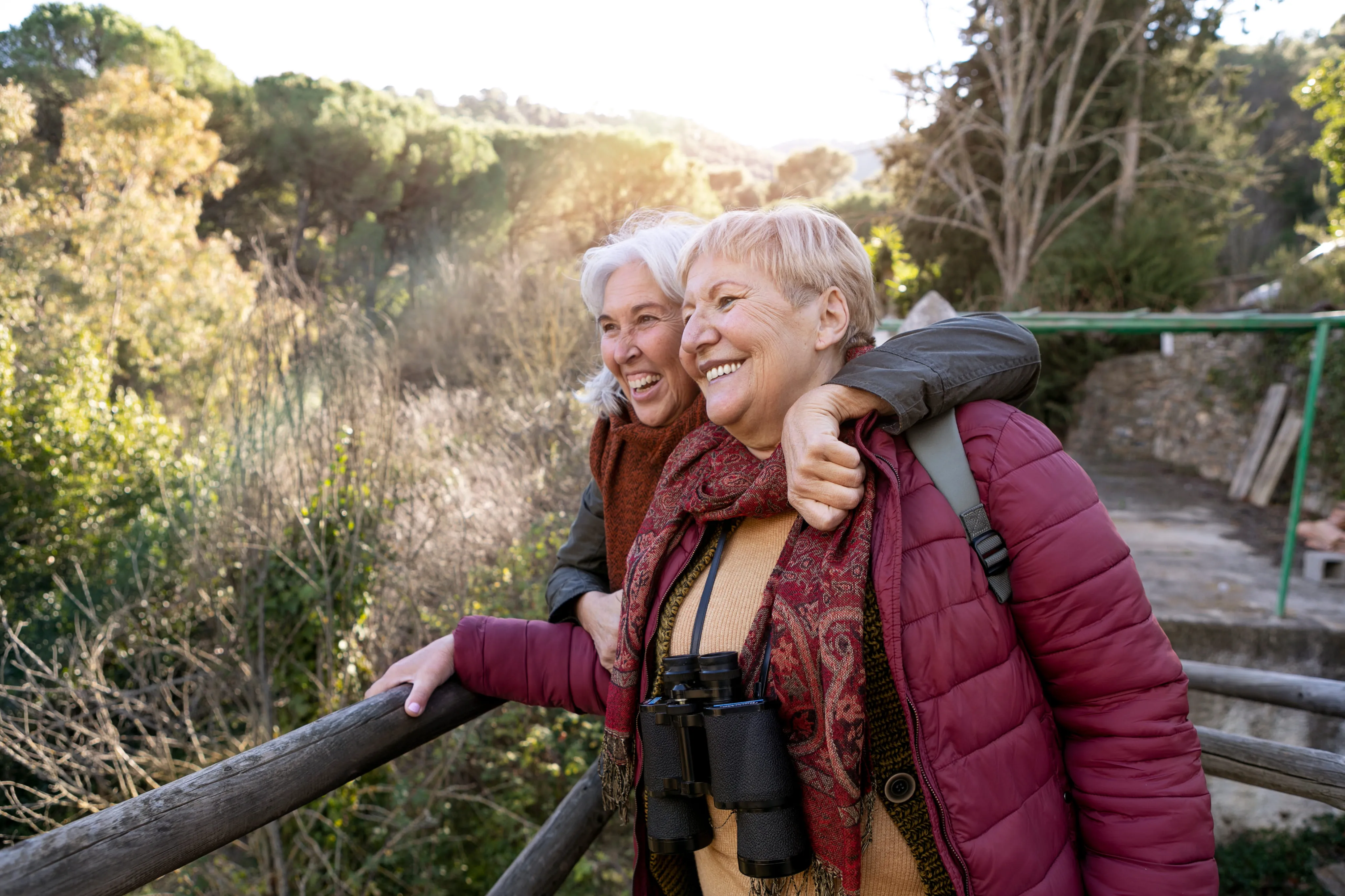 Two happy older women hugging and smiling outdoors near wooden railing and trees in sunlight.