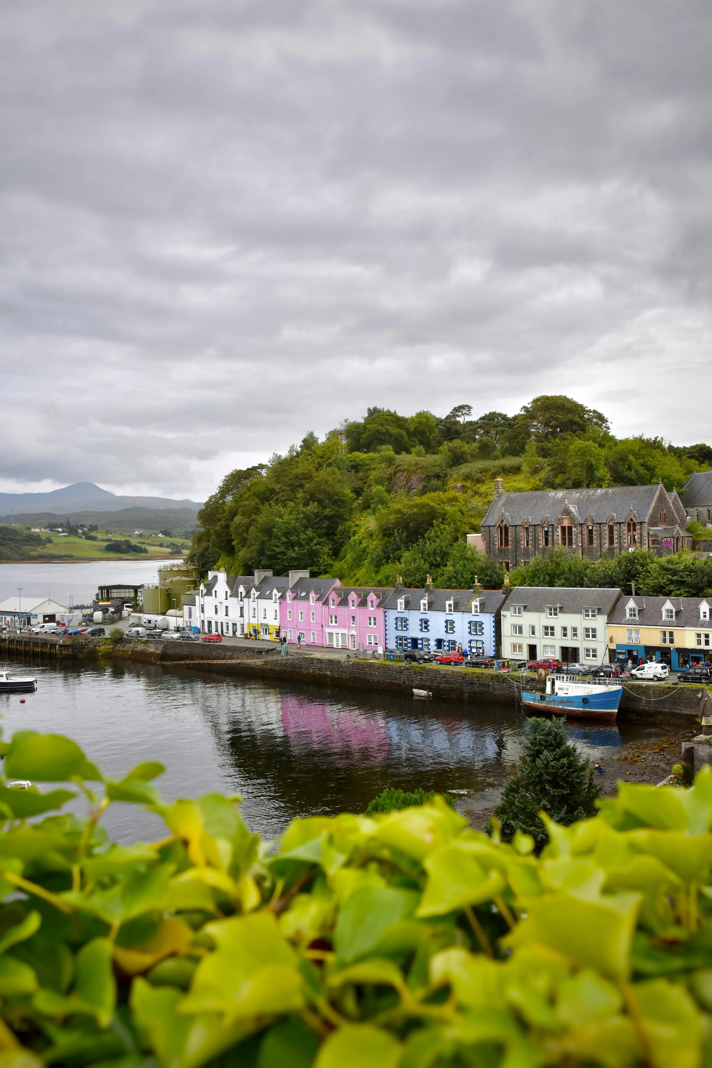 Colorful row of houses by a calm waterfront with boats and green hill behind under cloudy sky.