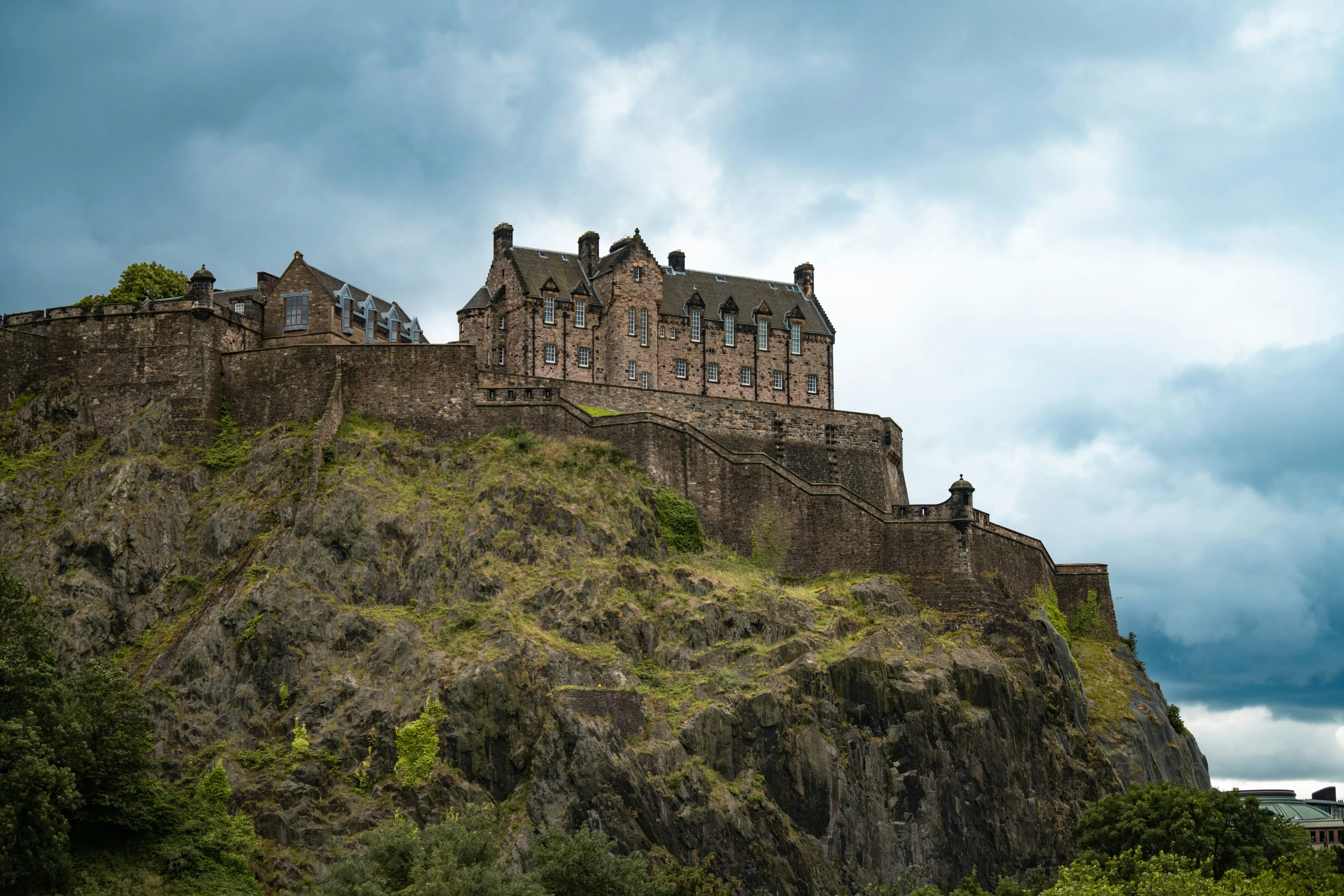 Historic stone castle atop a rocky hill under a cloudy sky, surrounded by green trees.