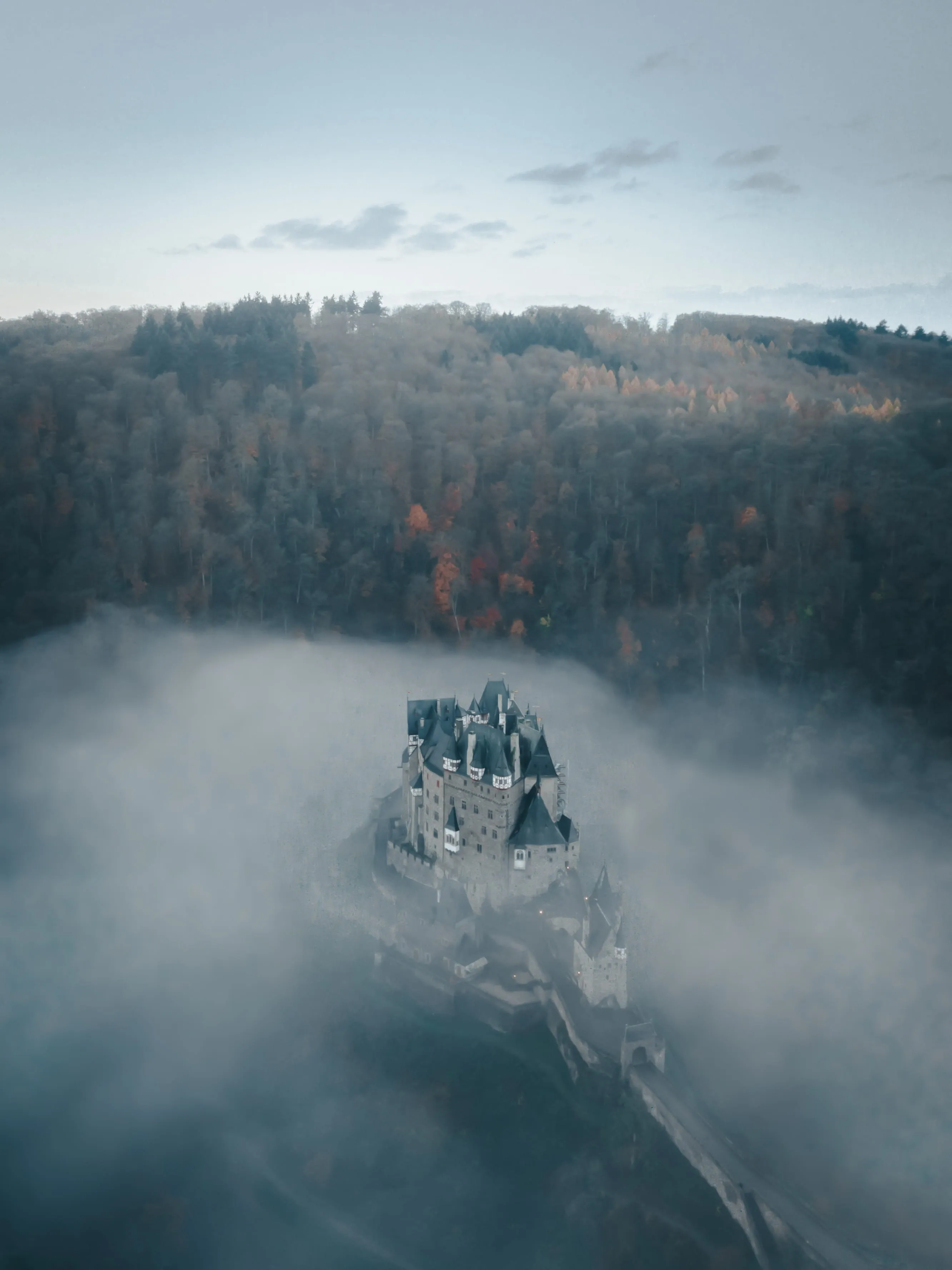 A castle surrounded by thick fog with a forest in the background under a cloudy sky.