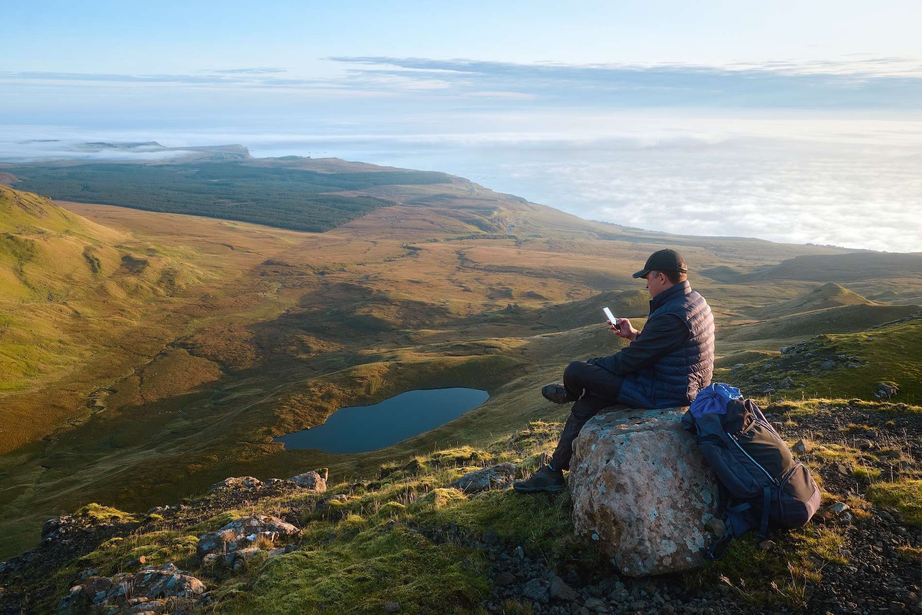 Man in outdoor gear sitting on rock overlooking valley and lake, using smartphone with backpack beside him.