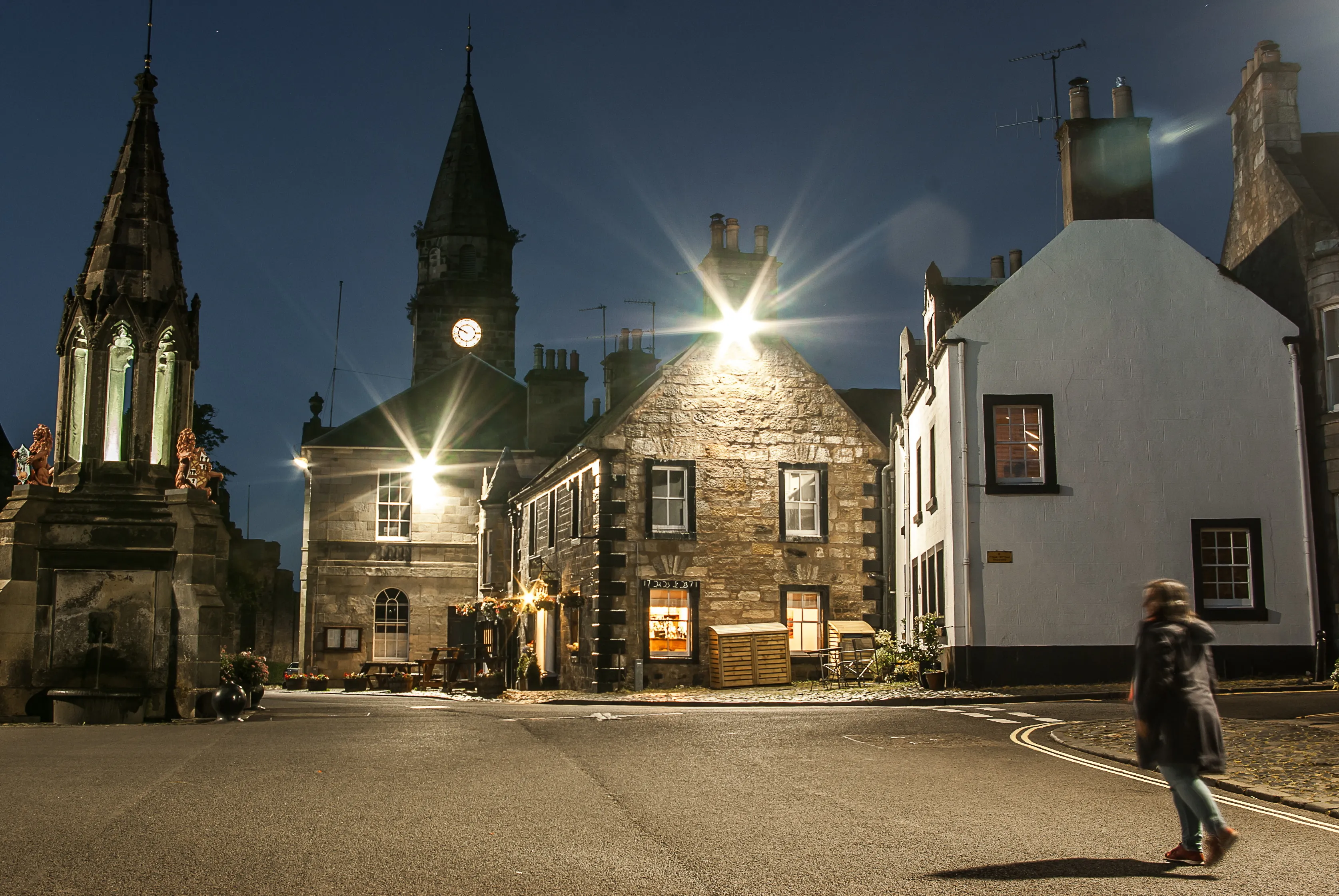A person walking near old stone buildings and a clock tower lit up at night on a quiet street.