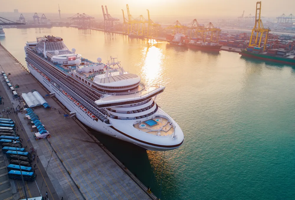 Large cruise ship docked at port with cranes and containers at sunset over calm water.