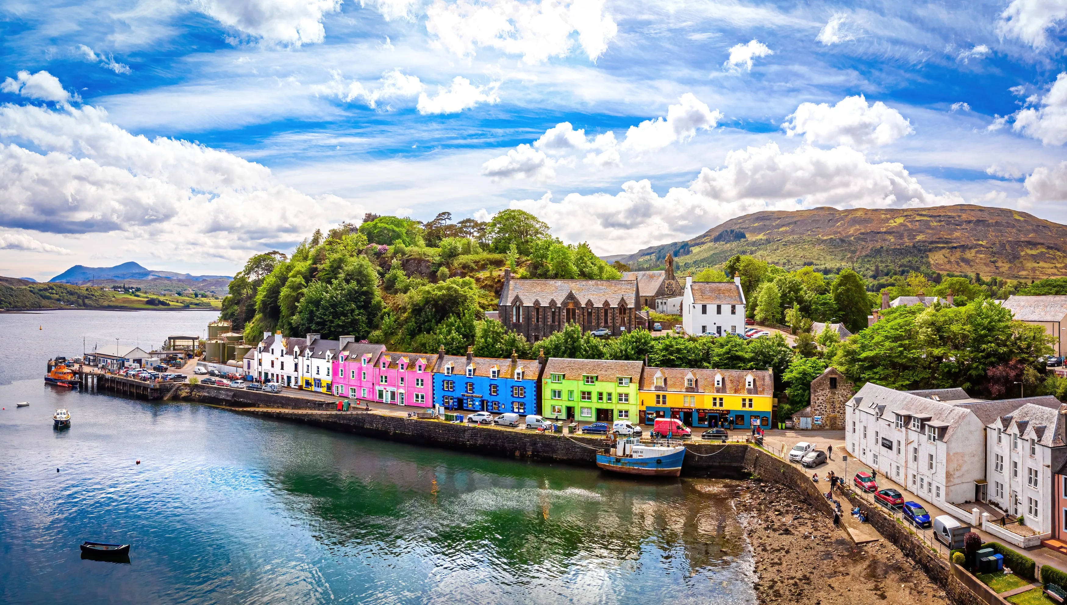 Colorful houses line a harbor with boats, backed by green hills and partly cloudy blue sky.