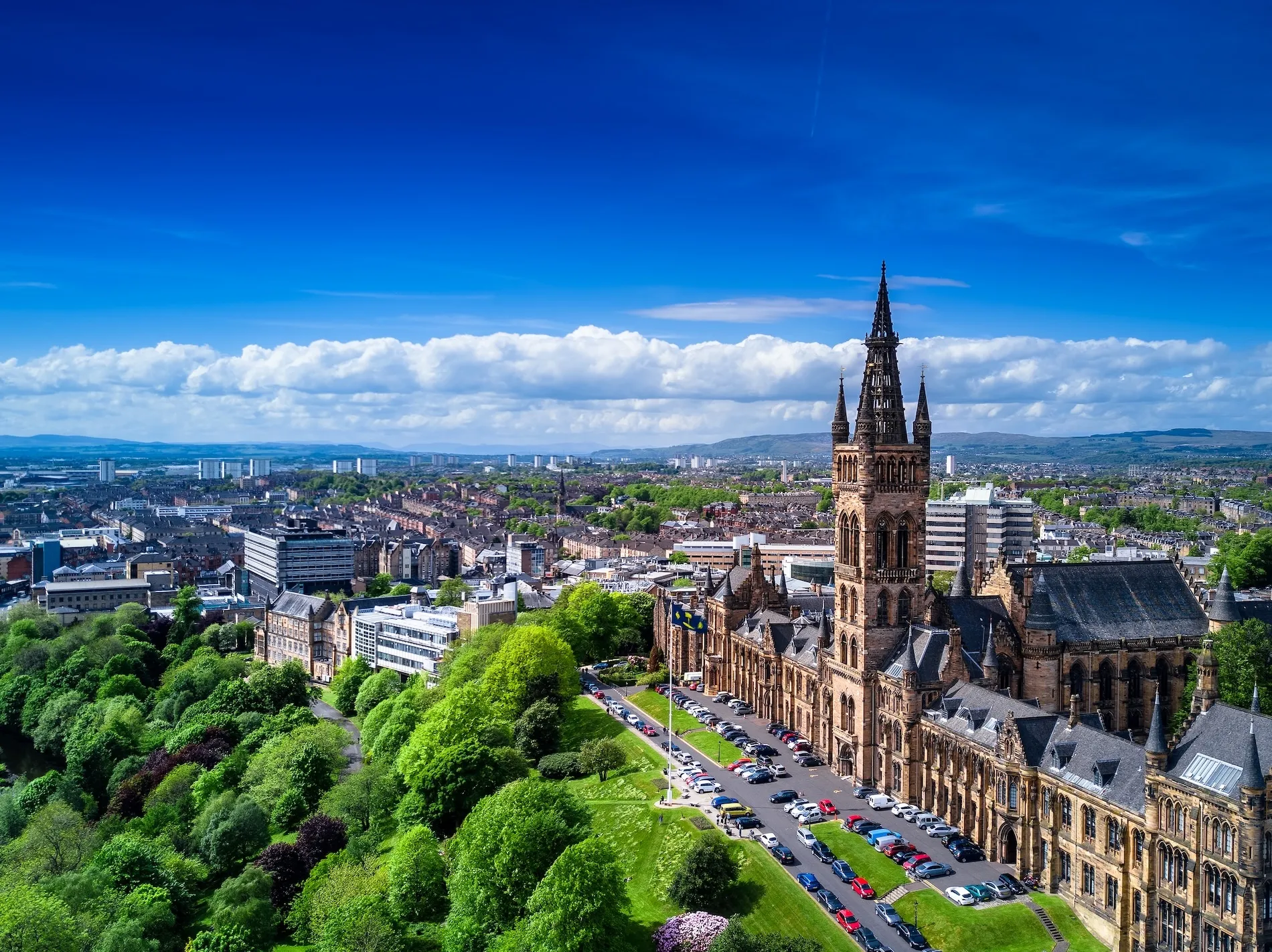 Glasgow University building with tall tower surrounded by green trees and cityscape under a blue sky.