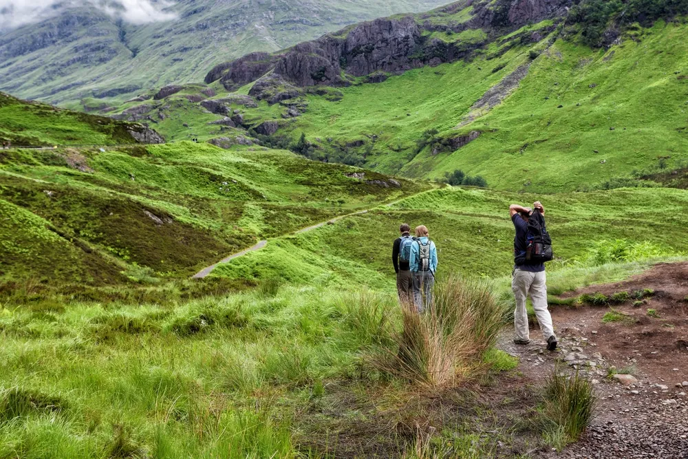 Three hikers with backpacks walking on a trail through green hills under cloudy skies.