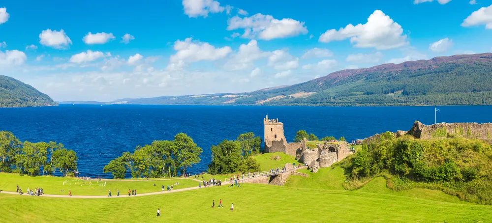 Ruins of a castle by a large blue lake with green hills and scattered clouds in a bright sky.