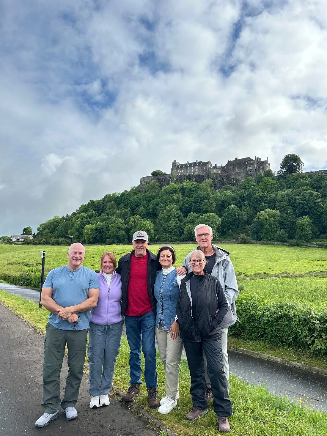 Six adults standing on a path in front of green hills and a castle under a cloudy sky.