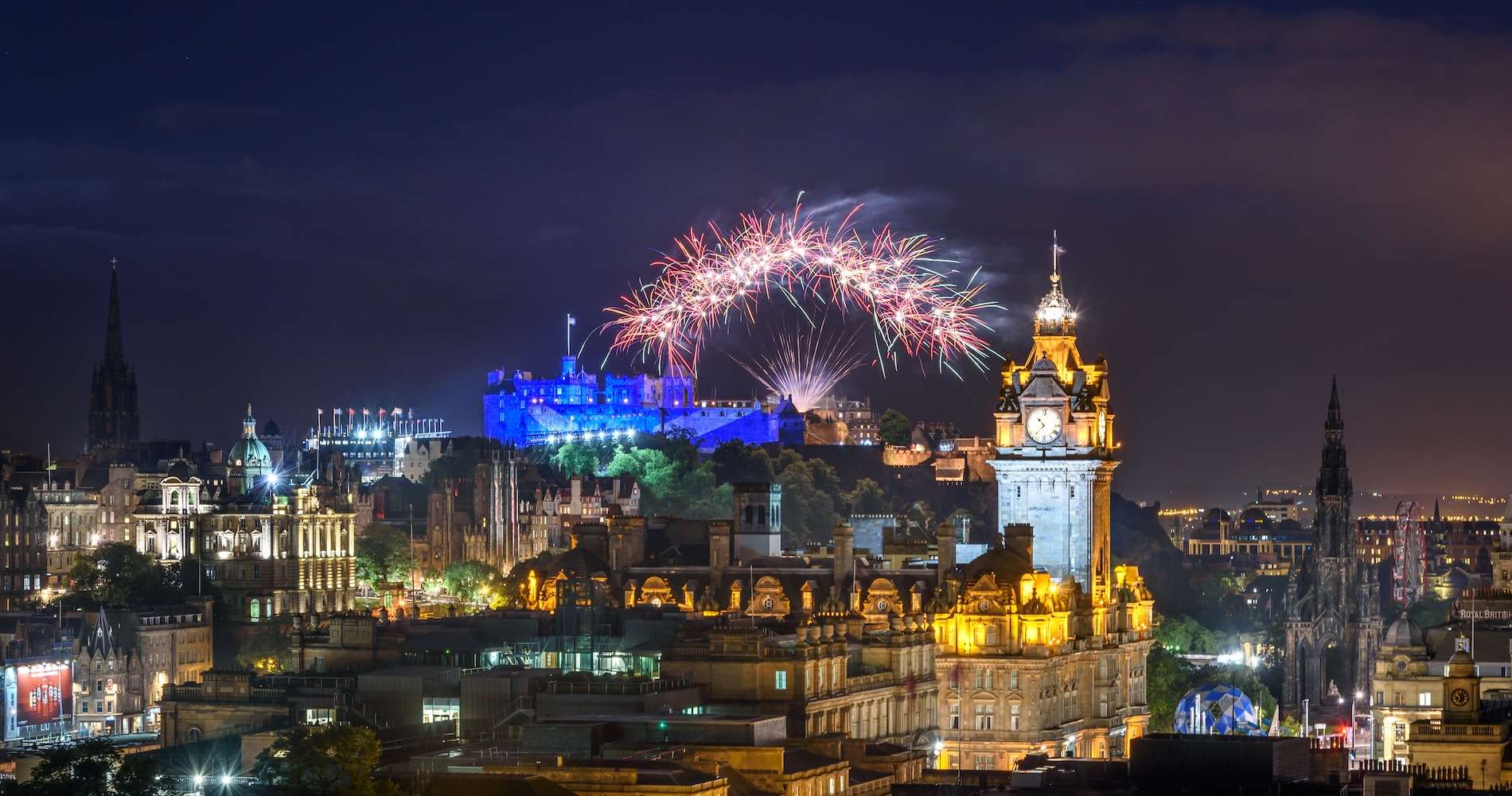 Fireworks over Edinburgh Castle illuminated in blue with the city skyline at night.