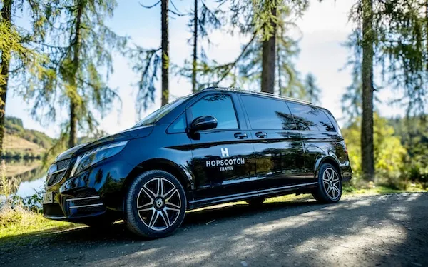 Black Hopscotch Travel van parked on a dirt road near trees and a lake in daylight.