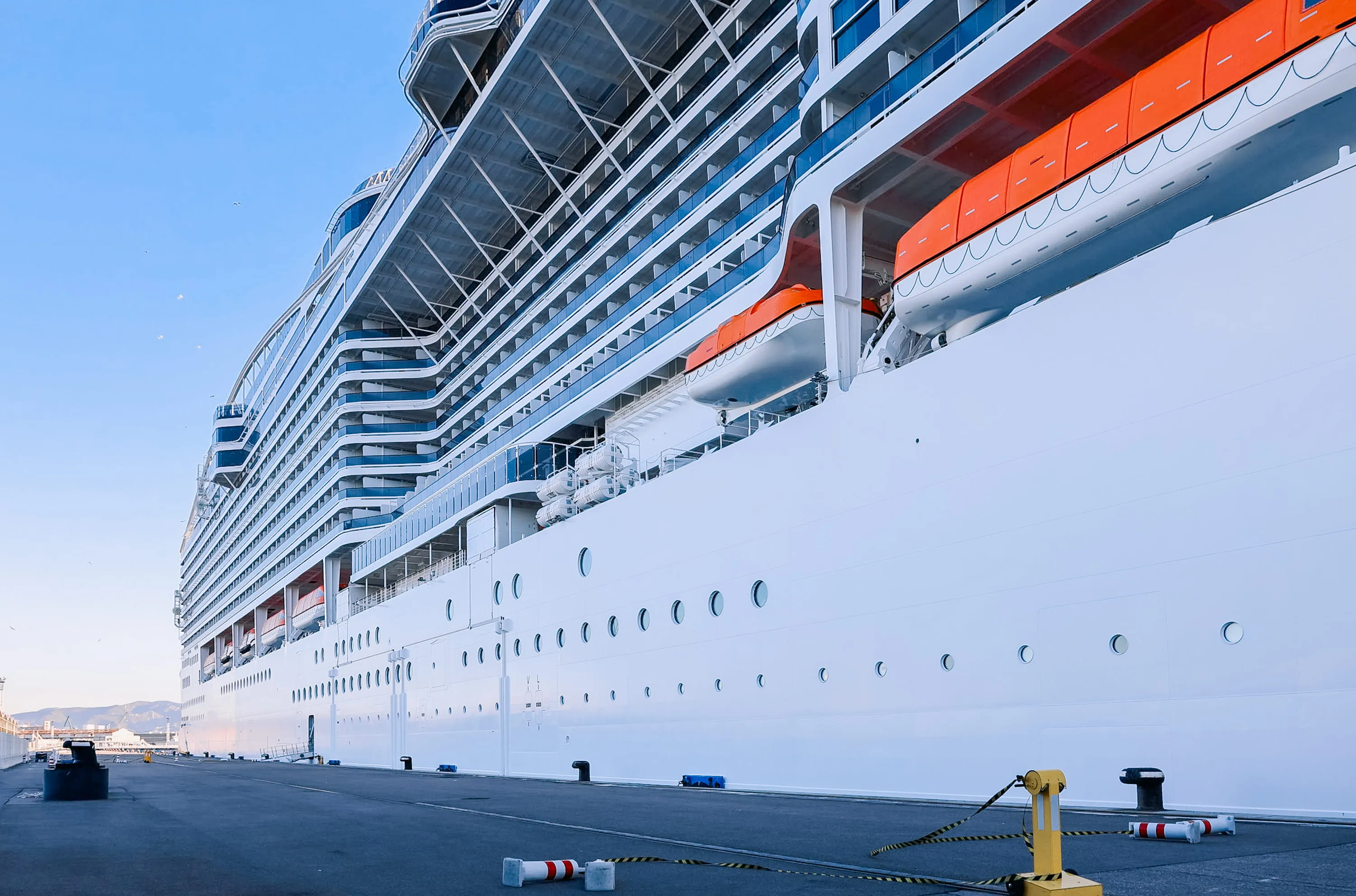 Side view of a large white cruise ship docked at a port with lifeboats and balconies visible.