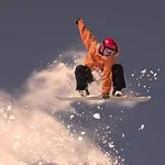 Snowboarder in orange jacket and red helmet mid-air against a blue sky.