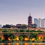 Traditional pagoda surrounded by trees with modern city buildings in the background at dusk.