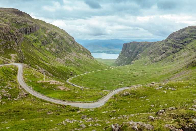 Winding single-track road through green mountains with a body of water visible in the distance.