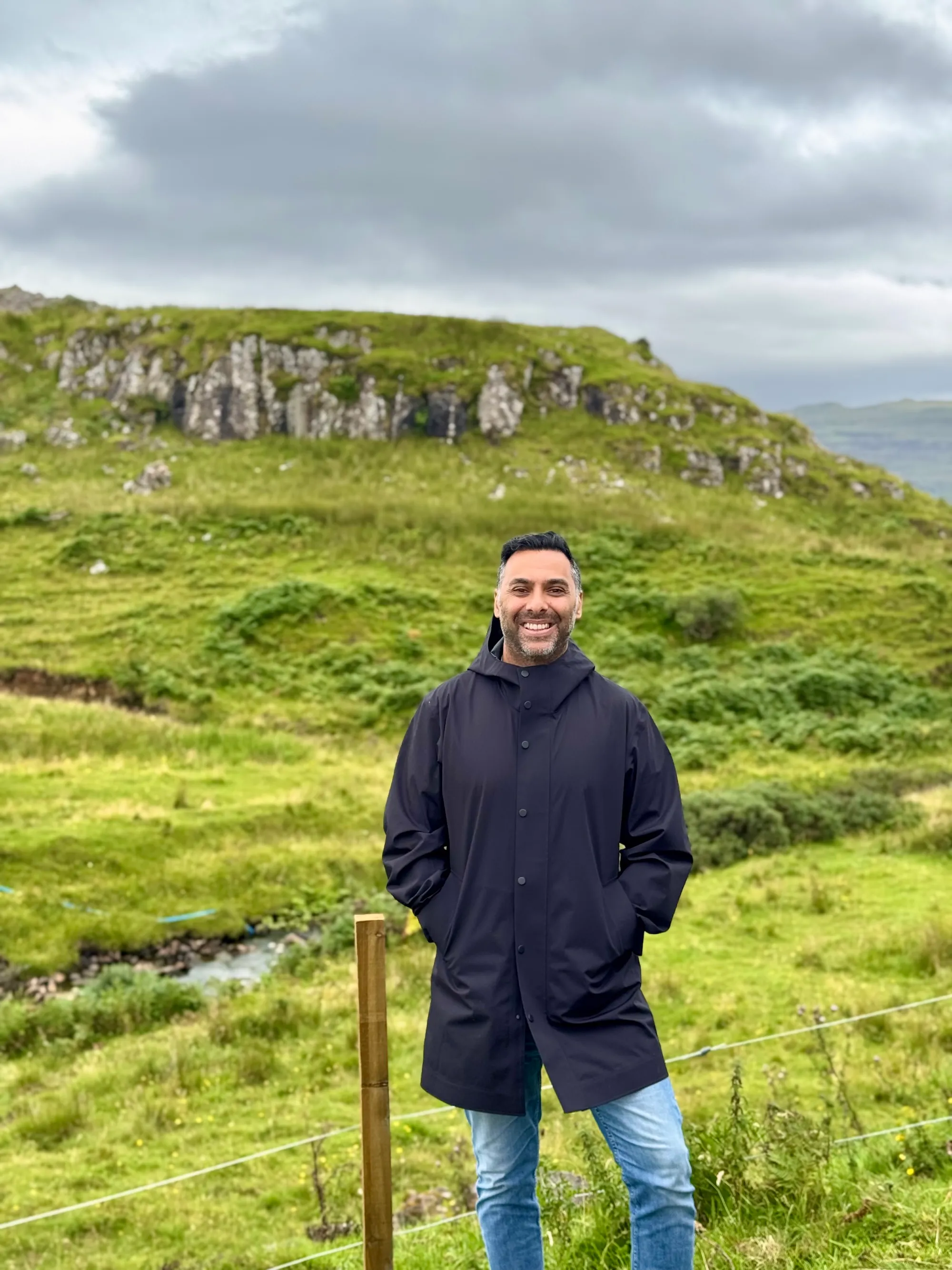 Smiling man in a black coat and jeans standing in a green grassy field with rocky hills behind him.