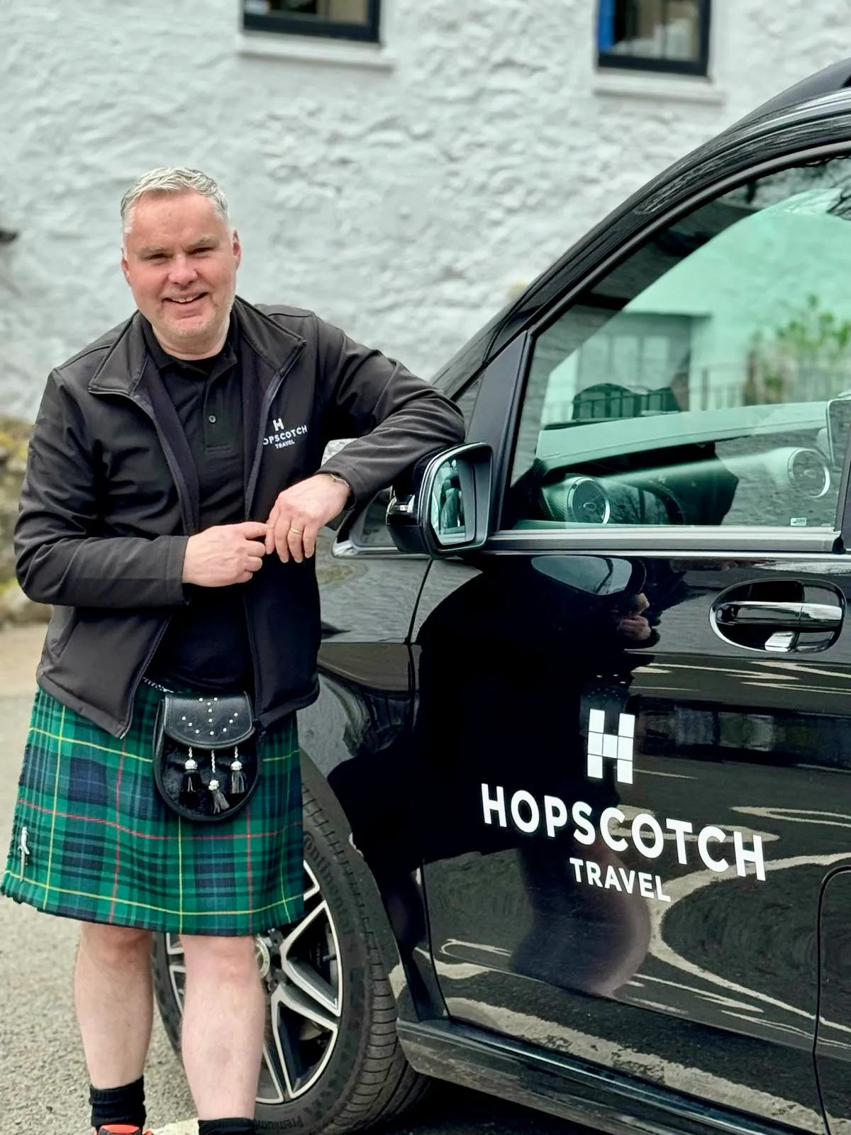 Smiling man in a green tartan kilt and jacket stands leaning on a black Hopscotch Travel van.