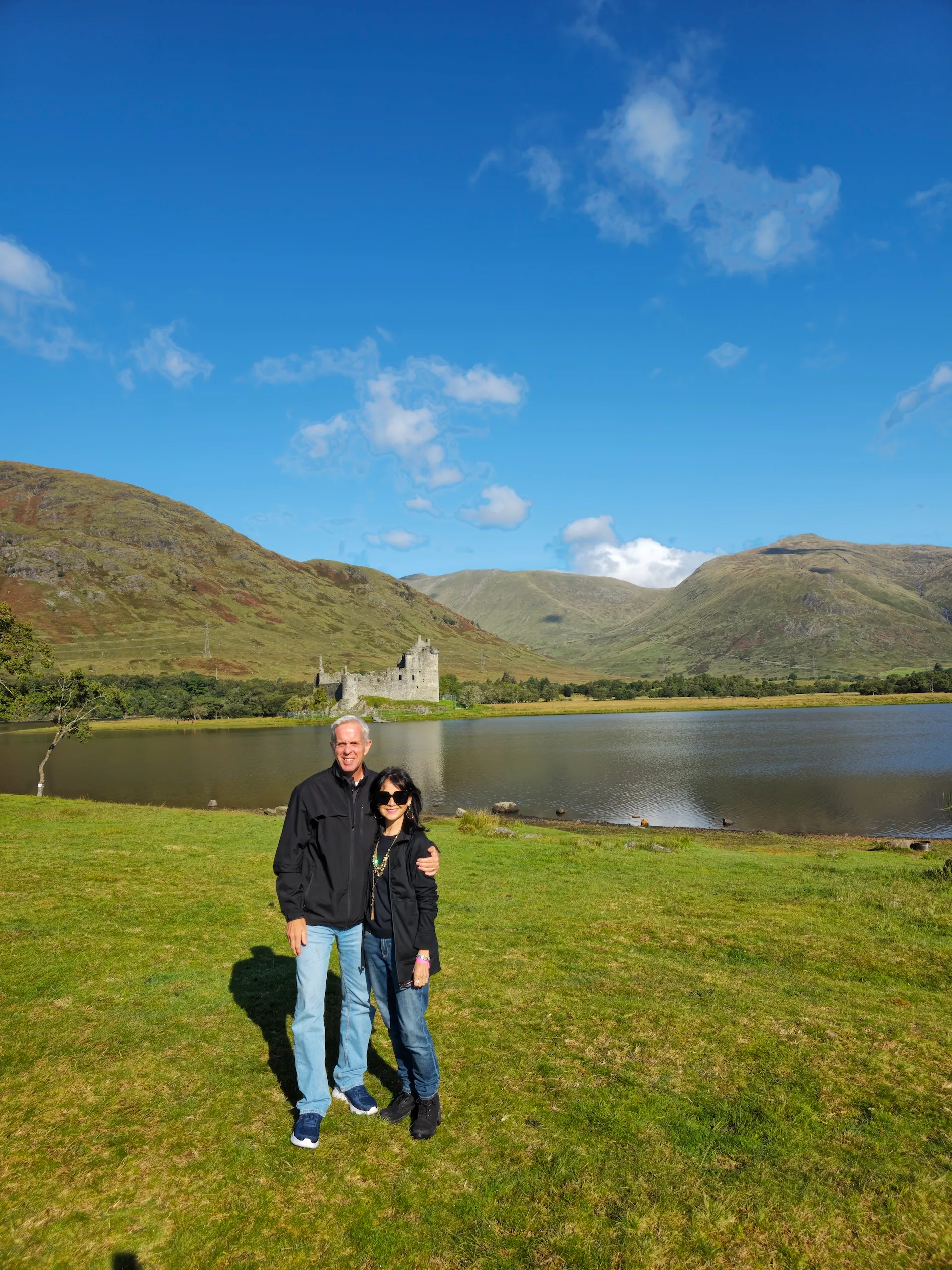 Couple standing on green grass near a lake with Kilchurn Castle and hills under a blue sky.