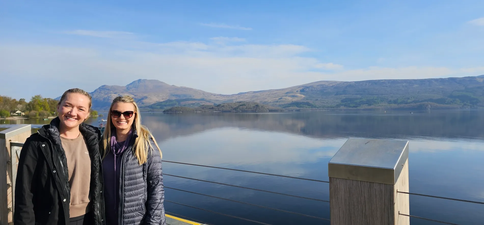 Two women smiling on a lakeside promenade with mountains and calm water in the background under blue sky.