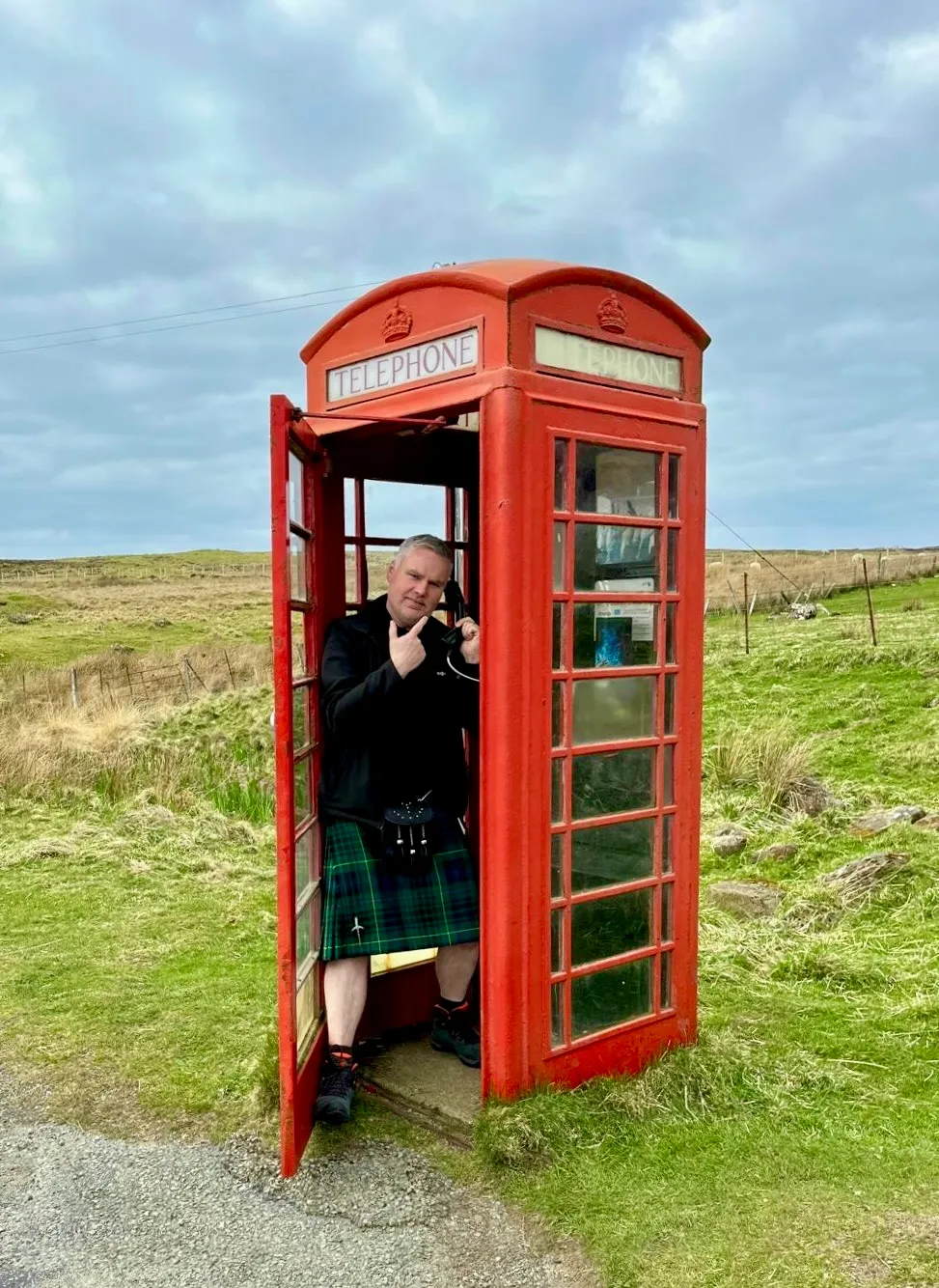 Man wearing a kilt inside a traditional red British telephone booth in a grassy rural area.