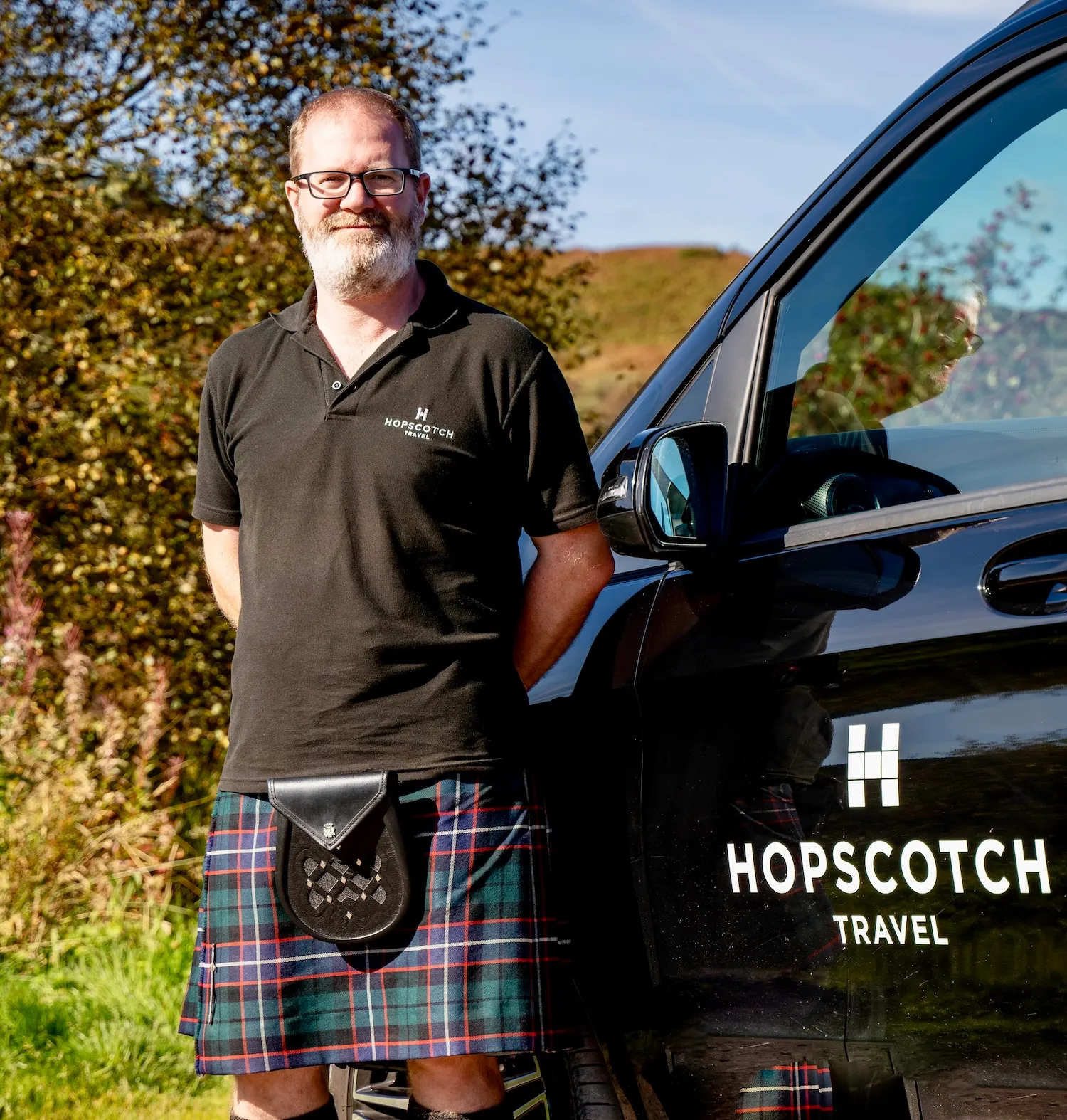 Bearded man in glasses, Hopscotch Travel shirt, and kilt stands beside black vehicle with company logo.
