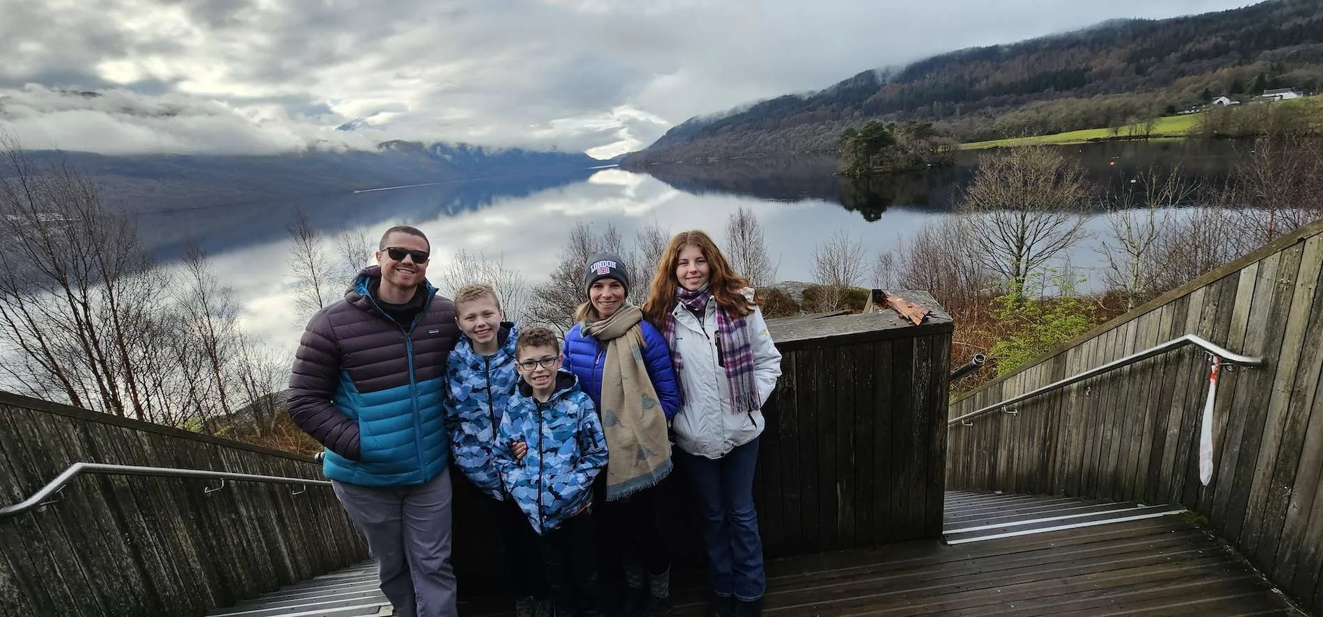 Five people standing on wooden stairs with misty lake and forested hills in the background.