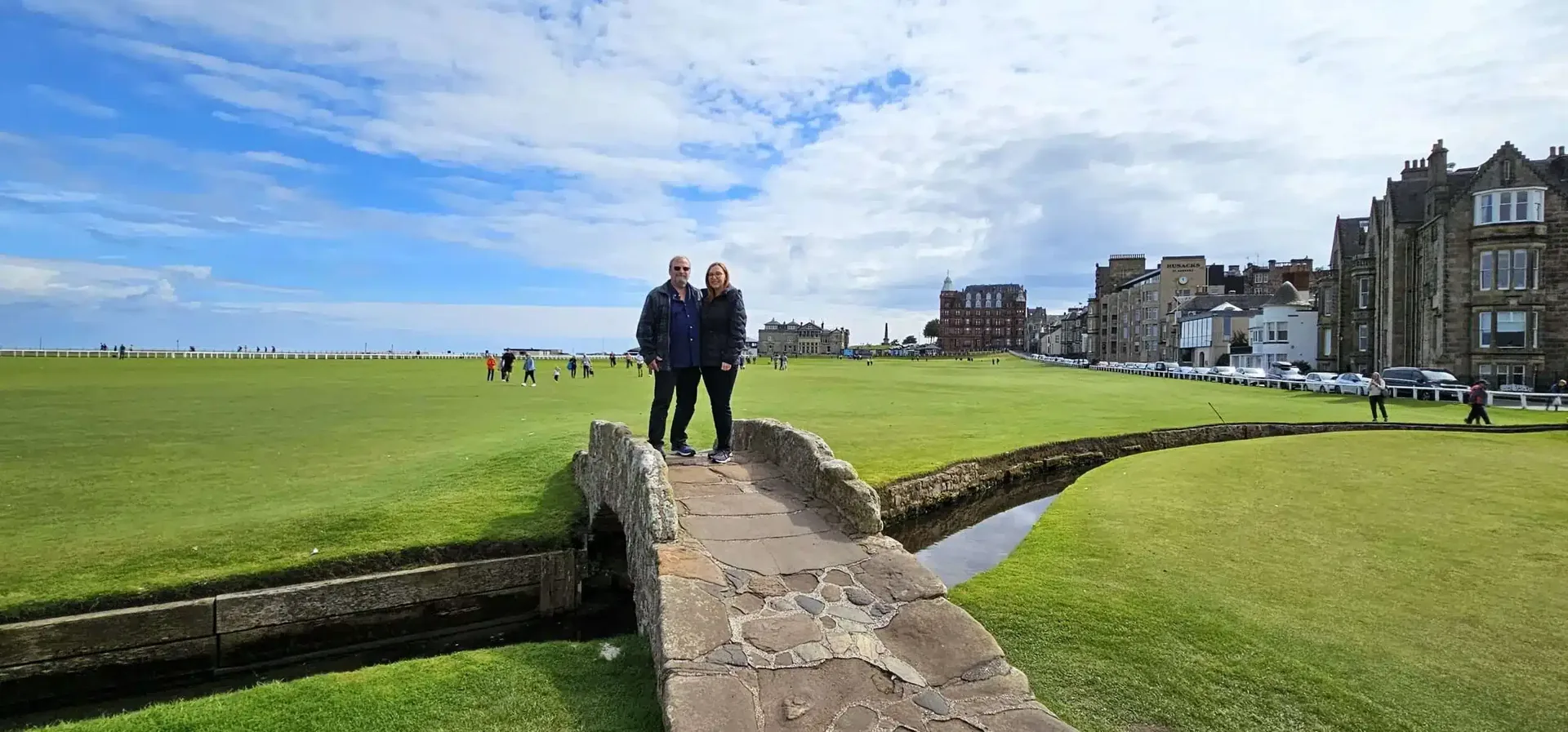 Couple standing on stone bridge over golf course water trench with buildings and blue sky in background.