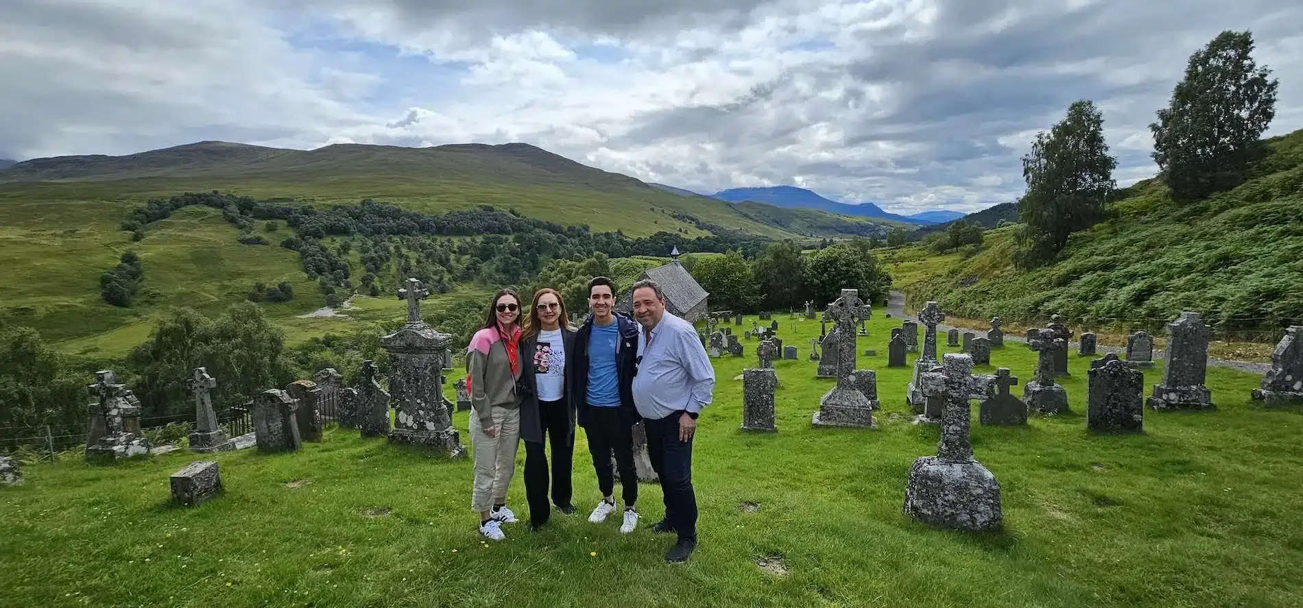 Four people standing together in a green cemetery with hills and cloudy sky in the background.