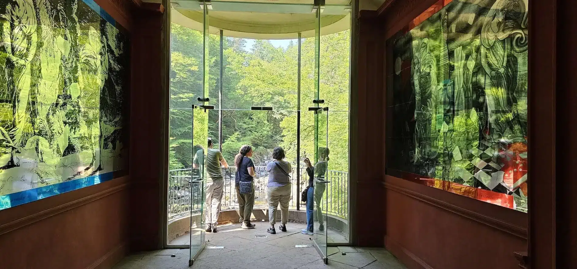 People standing on a balcony looking out at green trees through open glass doors inside a dim room.