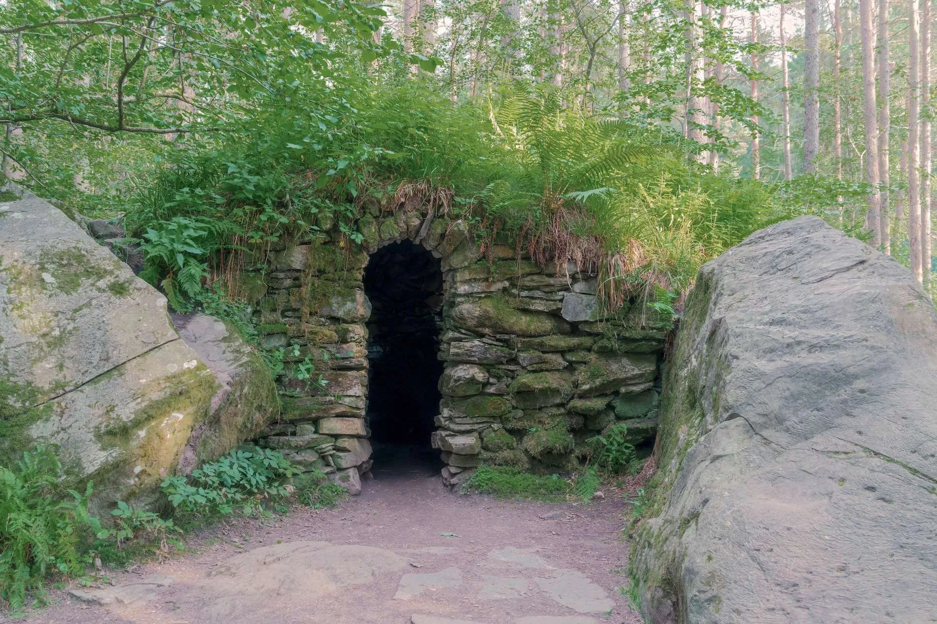 Stone arch entrance covered with moss and plants in a forest between two large rocks.