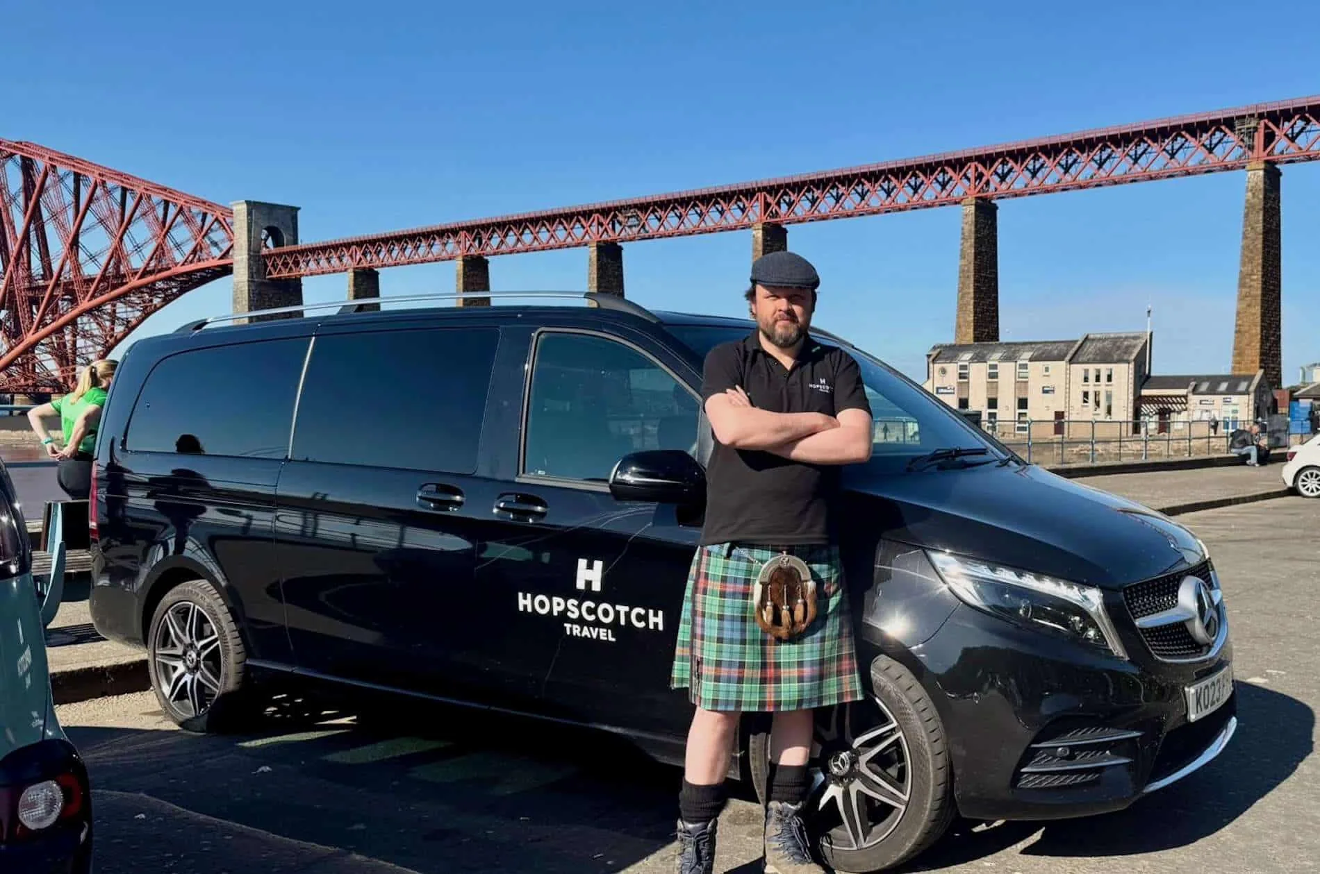 Man in black shirt and kilt standing with arms crossed by black Hopscotch Travel van near a large bridge.