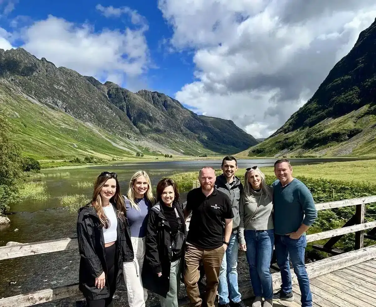 Group of six people standing on a wooden bridge with mountains and a lake in the background.