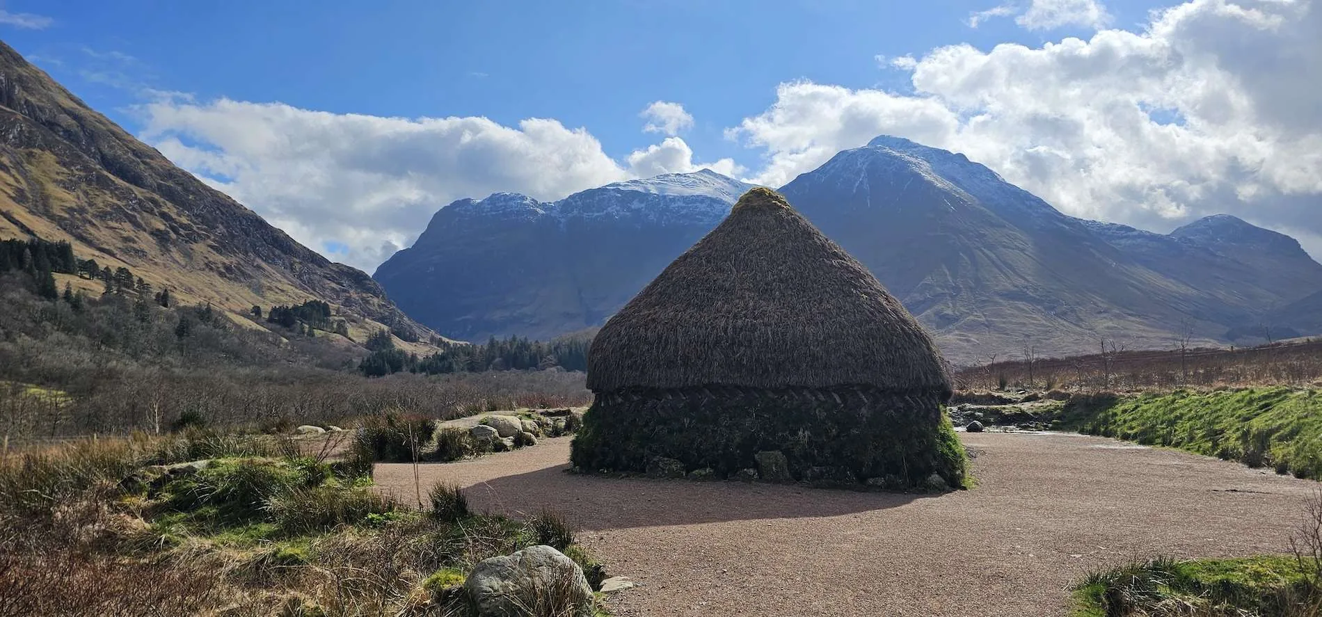 Thatched round hut with dark base in Glencoe valley with snow-capped mountains and blue sky.