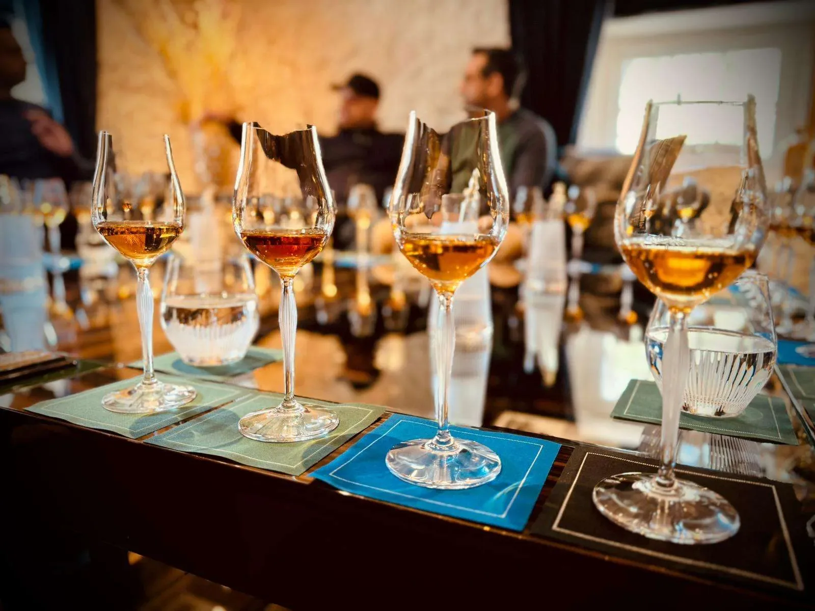 Four glasses of whisky with amber liquid on a table in a tasting setting with blurred people in background.